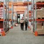 ALO Bringing 400+ Jobs to Southaven Distribution Center Workers walking down an aisle of stocked shelves inside a large warehouse, similar to a Southaven distribution center